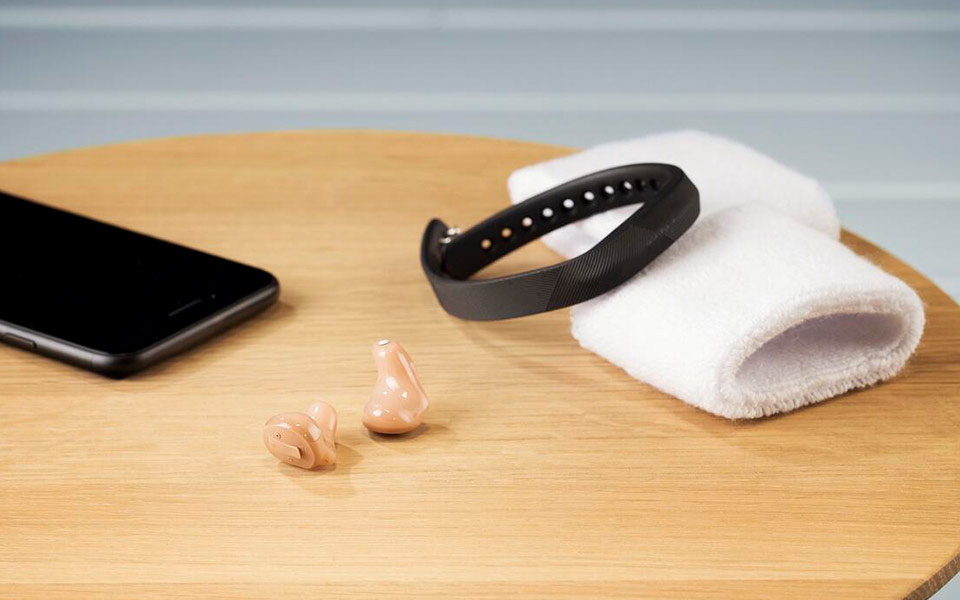 Zerena Im-Ohr Hörgeräte auf Tisch mit Schweißband, Smartband und Handy – symbolisiert den optimalen Einsatz im Fitnessstudio