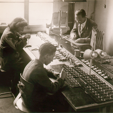 Vanha mustavalkoinen valokuva kolmesta työntekijästä istumassa pöydän ympärillä kokoamassa Bernafon-kuulokojeita, noin 1950- luvulla.