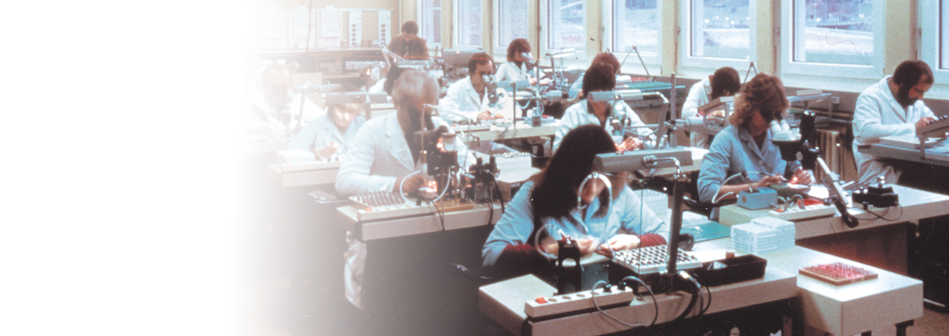 Gammalt foto av ett rum fullt av engagerade arbetare i laboratorierockar som arbetar med Bernafons hörapparatproduktion, 1989