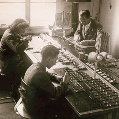 Vieja foto en blanco y negro de tres trabajadores sentados alrededor de una mesa armando audífonos Bernafon, alrededor de 1950