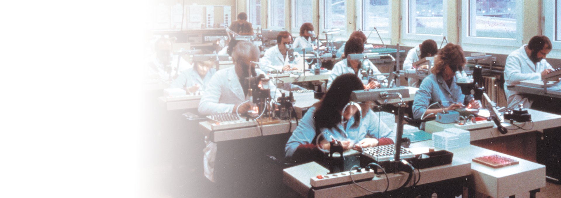 Vanha kuva, jossa on  huonetäynnä laboratoriotakkeissa olevia omistautuneita työntekijöitä, jotka työskentelevät Bernafonin kuulokojeiden valmistuksessa, 1989