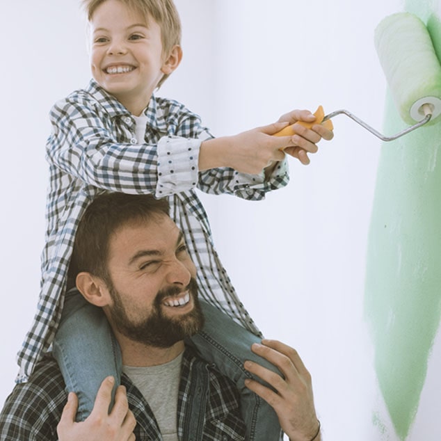 El padre lleva a su hijo con los audífonos detrás de la oreja Bernafon Leox Super Power | Ultra Power en sus hombros para pintar una pared.
