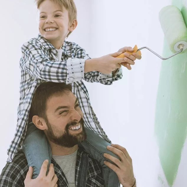 Father carries son with Bernafon Leox Super Power|Ultra Power behind-the-ear hearing aids on his shoulders to paint a wall. 