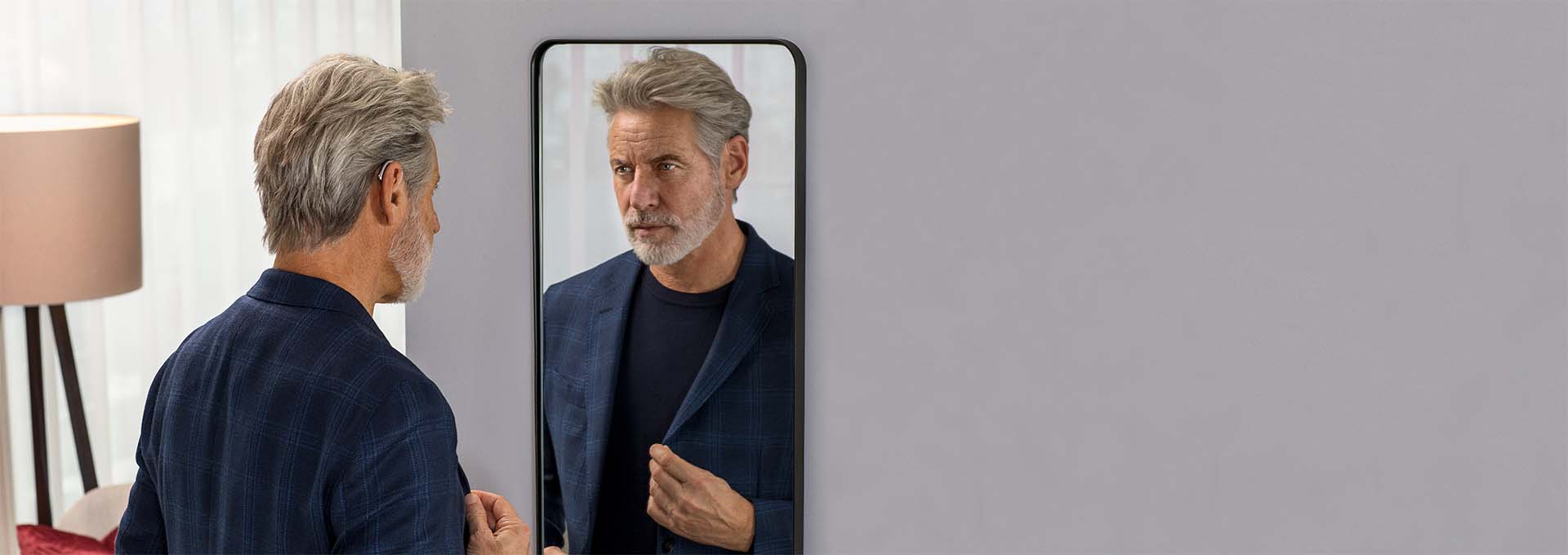 Hombre con audífonos miniBTE T R recargables de Bernafon de pie mirándose en un espejo, lleva un traje de chaqueta