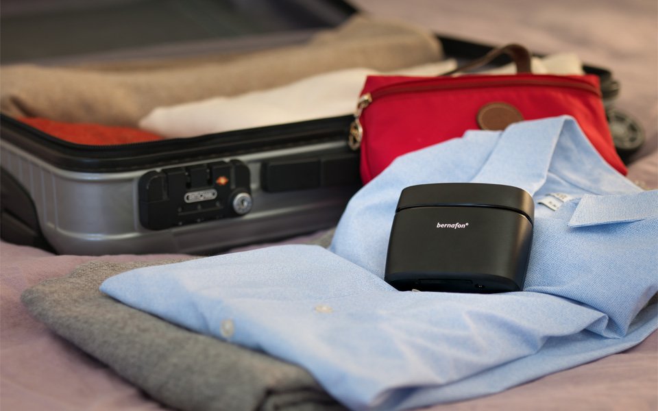 Charger Plus with Bernafon Alpha XT hearing aids laying next to an open suitcase on top of a blue shirt on the bed