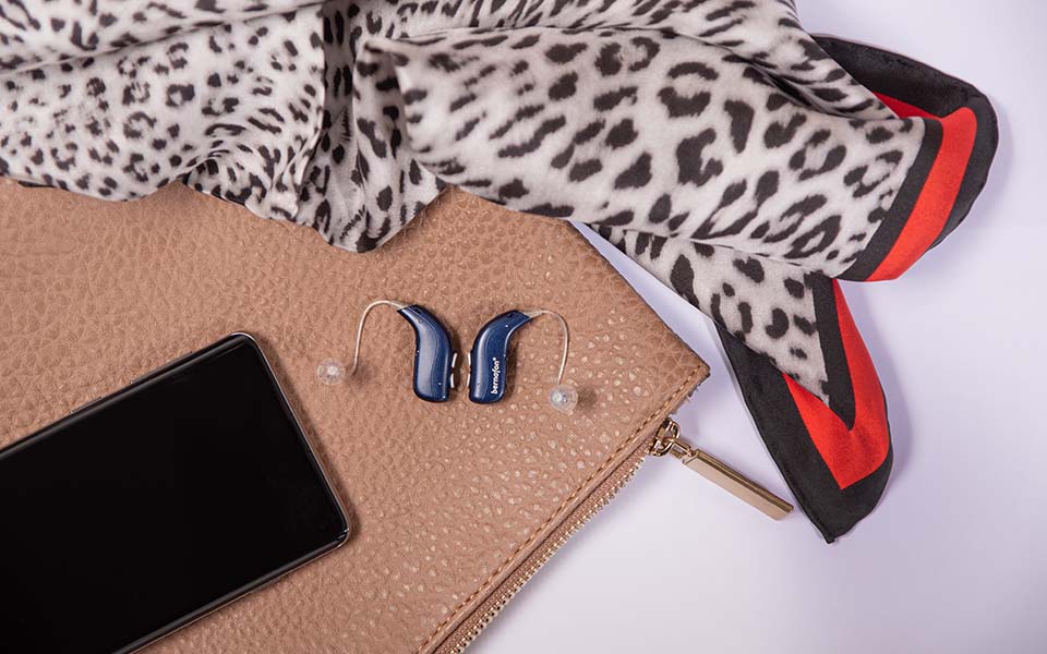 Midnight blue Bernafon Alpha rechargeable hearing aids sit on a purse next to a smartphone and animal print scarf