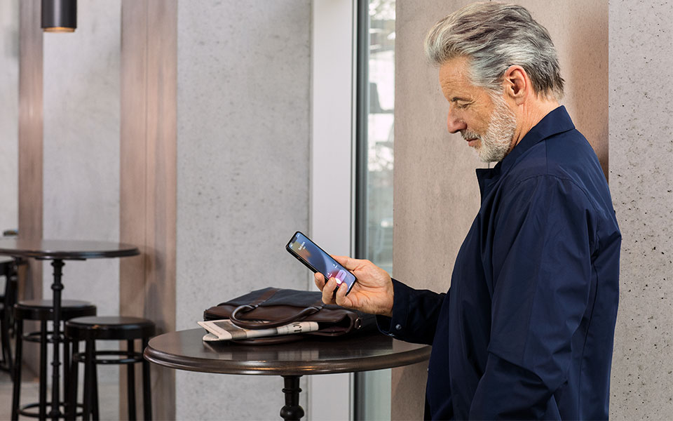 Hombre con audífonos miniBTE T R recargables de Bernafon de pie en una mesa alta en un restaurante con un iPhone en la mano