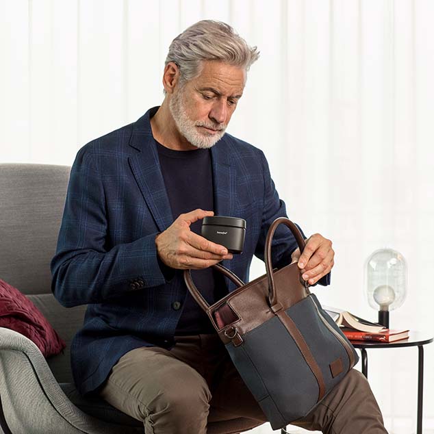 Hombre con audífonos miniBTE T R recargables de Bernafon sentado en la sala de estar y colocando el Charger Plus en su bolso