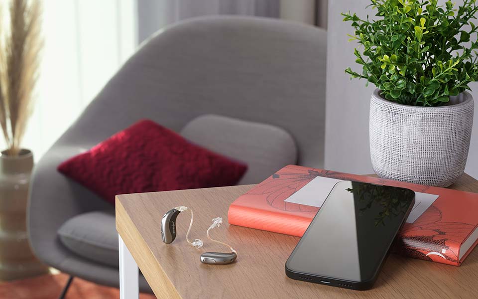 Dos audífonos miniBTE T R recargables de Bernafon, de color gris y negro, colocados sobre un escritorio en una sala de estar junto a un iPhone