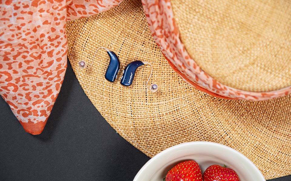 Paire d'appareils auditifs rechargeables Bernafon Alpha bleu foncé sur un chapeau de soleil d'été en paille avec une écharpe orange à imprimé animal. 