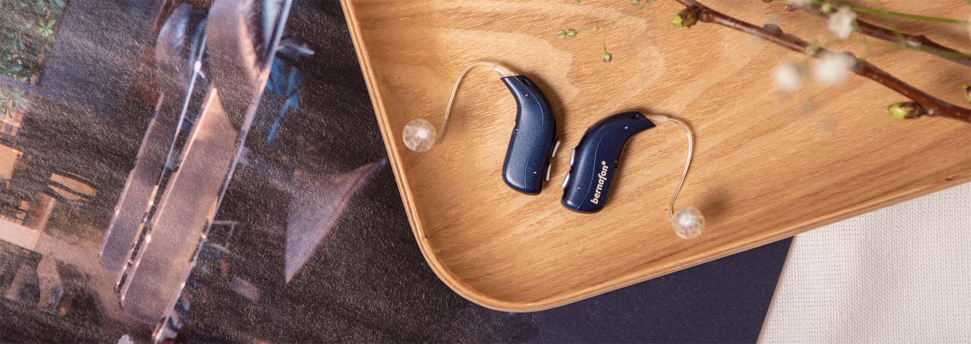 Paire d'appareils auditifs rechargeables Bernafon Alpha bleu foncé sur une planche de bois avec un magazine et des fleurs de printemps blanches 