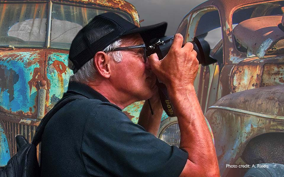 Utente di apparecchi acustici Bernafon Alpha (uomo) visto di lato mentre scatta una foto con la sua macchina fotografica in un vecchio cimitero di automobili