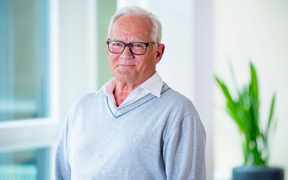 Utilisateur d'une aide auditive Bernafon Alpha (homme portant une chemise blanche et un pull bleu à col en V) debout et souriant à la caméra