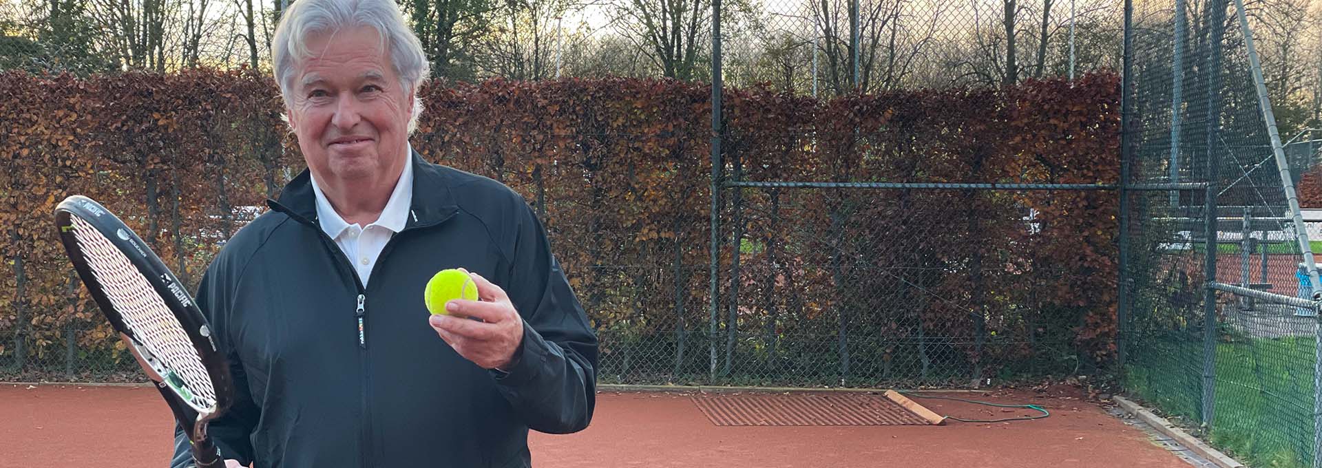Bernafon Alpha -kuulokojeen käyttäjä (mies) tennispallo ja maila kädessä kentällä katselee kameraan valmiina pelaamaan.
