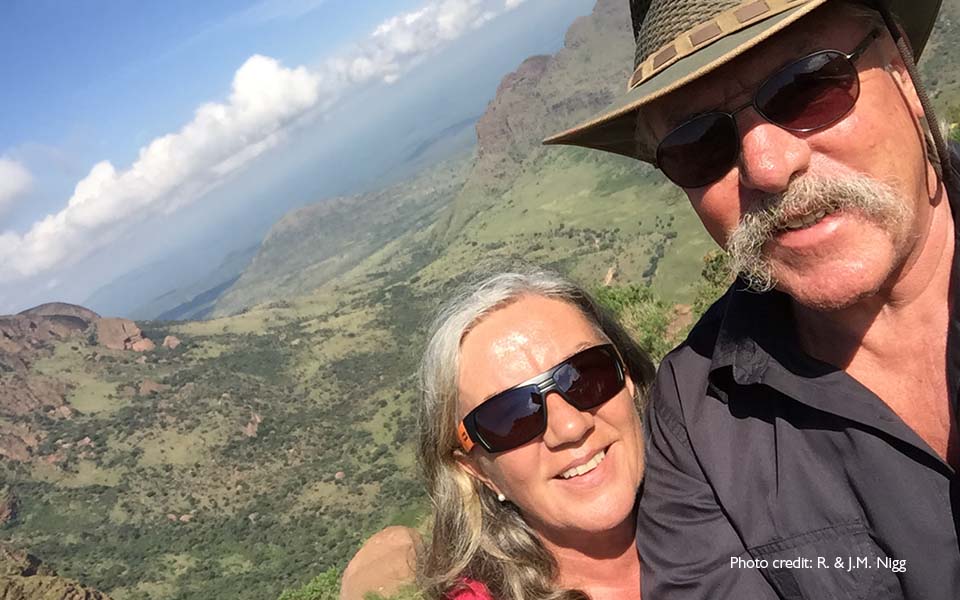 Güneş gözlüklü Bernafon Alpha işitme cihazı kullanıcıları (erkek ve kadın) arkadaki manzarayla birlikte bir tatil selfiesi çekiyor