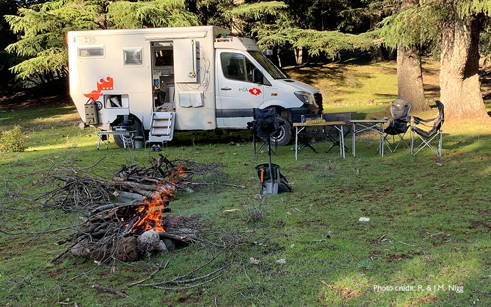 Camper van Bernafon Alpha hoortoesteldragers geparkeerd in een bos met kampvuur en in de namiddag schaduw op het gras