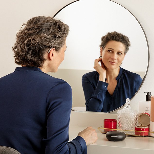 Woman wearing Bernafon Alpha rechargeable hearing aids sits in front of a mirror at a make-up table with hearing aid charger