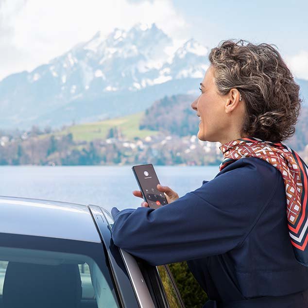 Frau trägt wiederaufladbare Bernafon Alpha Hörgeräte, steht an ihrem Auto, schaut auf einen Schweizer See und telefoniert dabei über ihr Smartphone.
