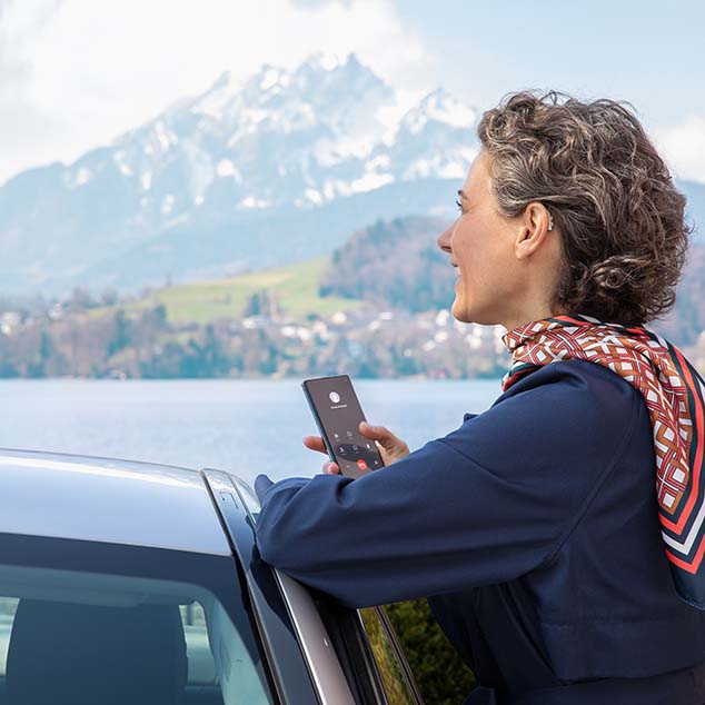 Woman wearing Bernafon Alpha rechargeable hearing aids stands by car at a Swiss lake and streams phone call from smartphone