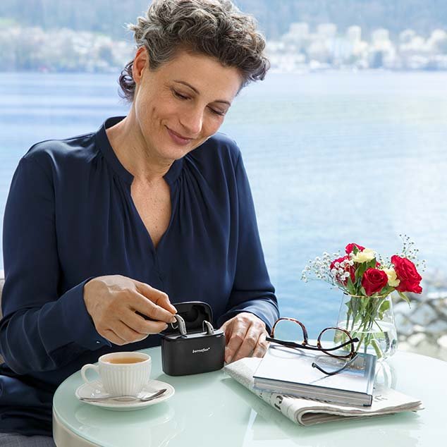 Frau sitzt an einem Tisch, im Hintergrund ein Schweizer See, sie entnimmt gerade ihre wiederaufladbaren Bernafon Alpha Hörgeräte aus der mobilen Ladestation Plus.