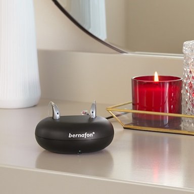 Aides auditives rechargeables Bernafon Alpha dans un chargeur sur une table de maquillage avec un miroir et un vase de fleurs séchées.
