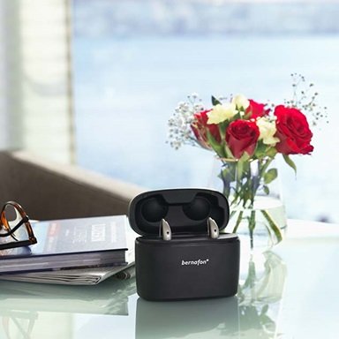 Aides auditives Bernafon Alpha rechargeables dans un chargeur portable Charger Plus sur une table en verre avec des fleurs rouges, un livre et des lunettes.