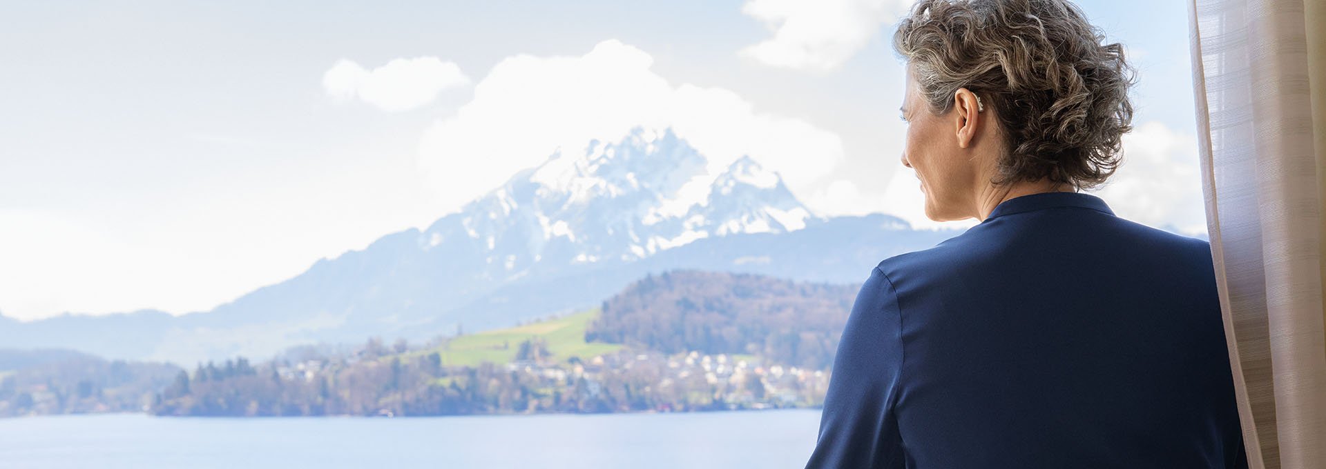 Mujer vistiendo audífonos recargables Bernafon Alpha disfruta de vistas al lago suizo y a la montaña desde la ventana del hotel