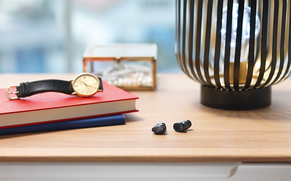 Two black Bernafon Alpha wireless ITC hearing aids laying on a desk next to two books, a watch, a jewelry box and a lamp