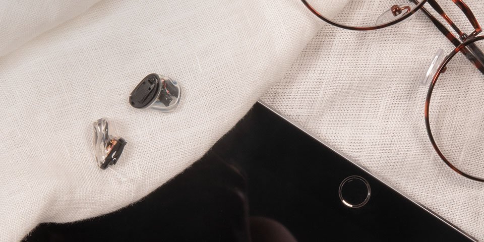 De zwarte en transparante kleinste in het oor hoortoestellen liggen op een wit laken naast een bril en tablet.