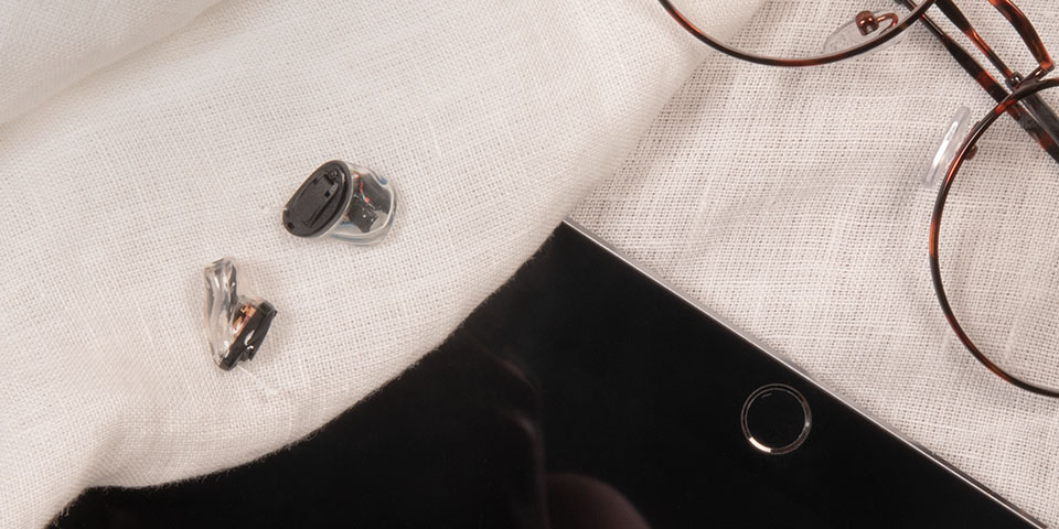 Los audífonos negros y transparentes más pequeños en el oído que se encuentran en una sábana blanca junto a gafas y tablets.