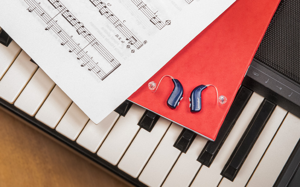 Los audífonos recargables Bernafon Alpha de color azul noche se encuentran sobre un cuaderno rojo en un piano junto a una hoja de notas musicales