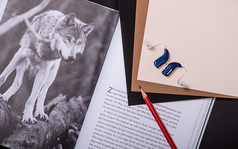 Audífonos recargables Bernafon Alpha azul noche colocados sobre un libro con la imagen de un lobo junto a un lápiz rojo