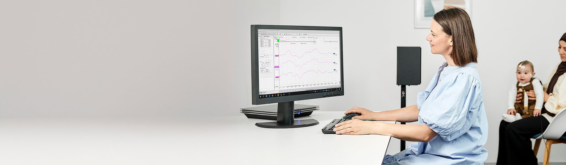 Niemowlę siedzące z mamą na krześle, podczas gdy protetyk słuchu wykonuje u niego test Aided Cortical.