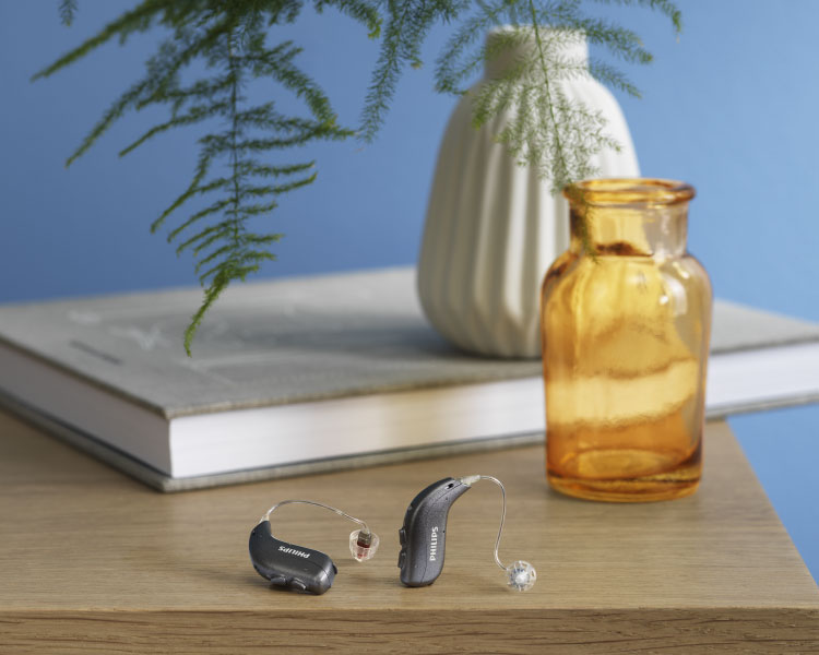 Philips HearLink non-rechargeable hearing aids placed on a wooden table, with a magazine, glasses and yellow plant pot