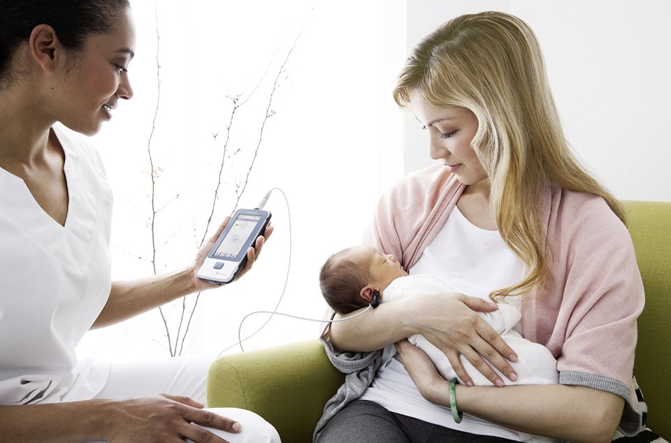 Audiologist testing a baby's hearing with an easyScreen ABR and OAE test device, while the mother holds the sleeping baby tight