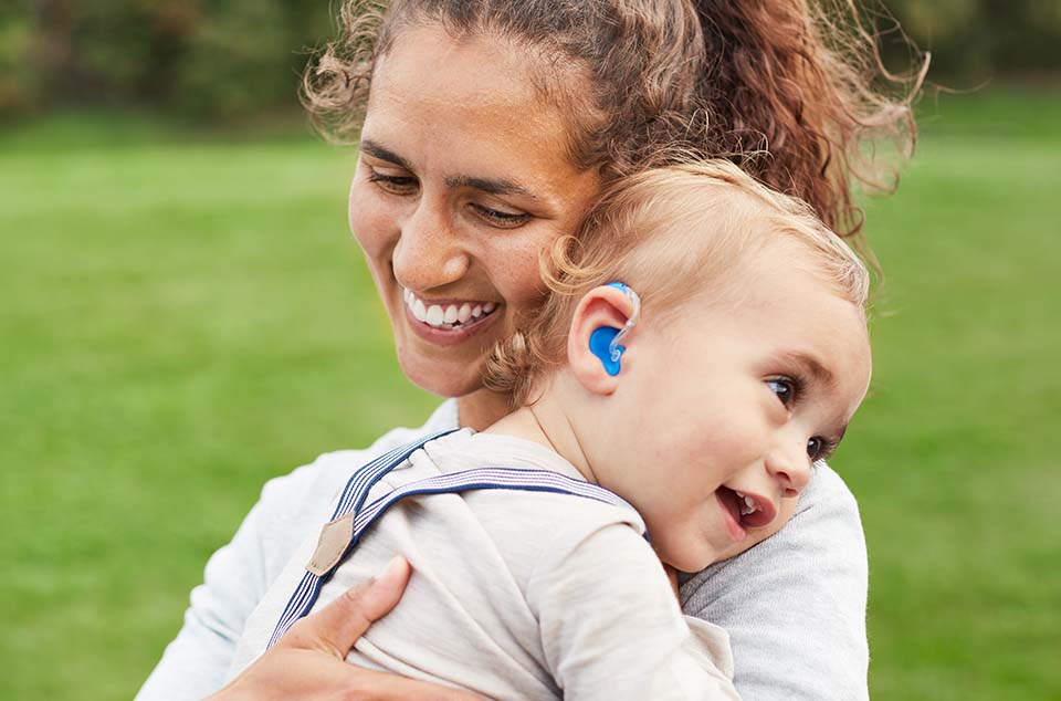 maman et son bébé avec un appareil auditif bleu riant aux éclats