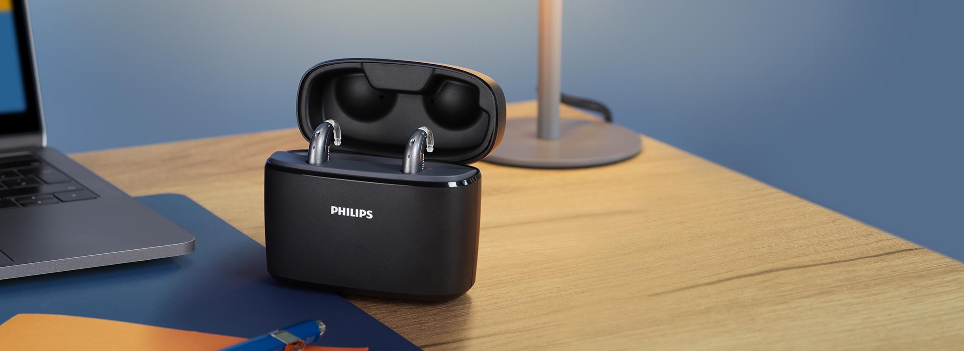 Philips HearLink rechargable hearing aids sit inside the portable Charger Plus, which lays on a home work desk next to a laptop, lamp, notebook and pen