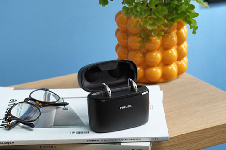 Audífonos recargables Philips HearLink en el cargador Plus de viaje colocados en una mesa de madera, con una revista, gafas y una maceta amarilla.