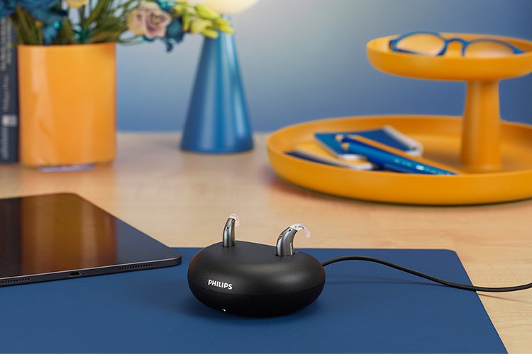 Audífonos Philips HearLink en el formato mini retroaricular (miniBTE T R) se cargan en el cargador de sobremesa en un escritorio de madera junto a una lámpara, un iPad, libros, flores y un organizador de escritorio