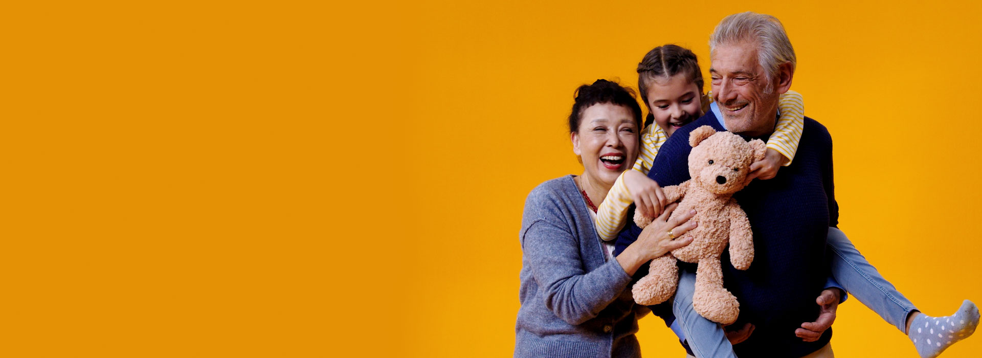 A granddaughter holds a teddy bear while sitting on the back of her grandfather who wears Philips HearLink hearing aids. Her mother also reaches out to hold her daughter.