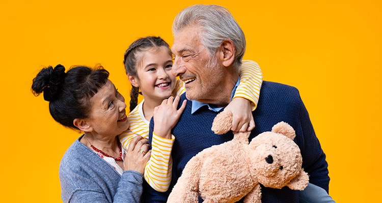 A granddaughter holds a teddy bear while sitting on the back of her grandfather who wears Philips HearLink hearing aids. Her mother also reaches out to hold her daughter.