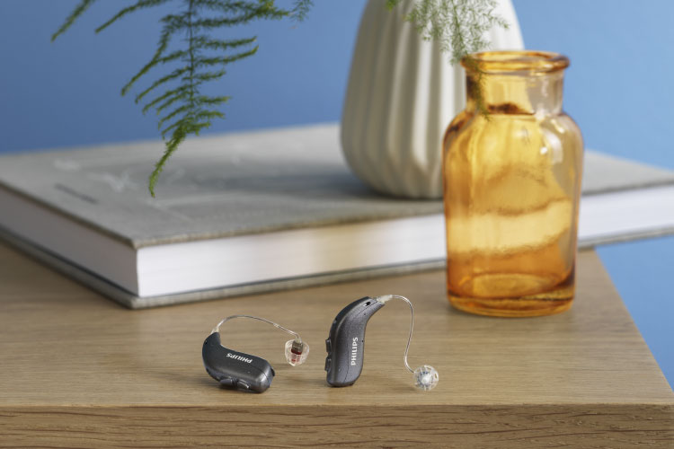 Appareils auditifs non rechargeables Philips HearLink placées sur une table en bois, avec un magazine, des lunettes et un pot de plantes jaunes.