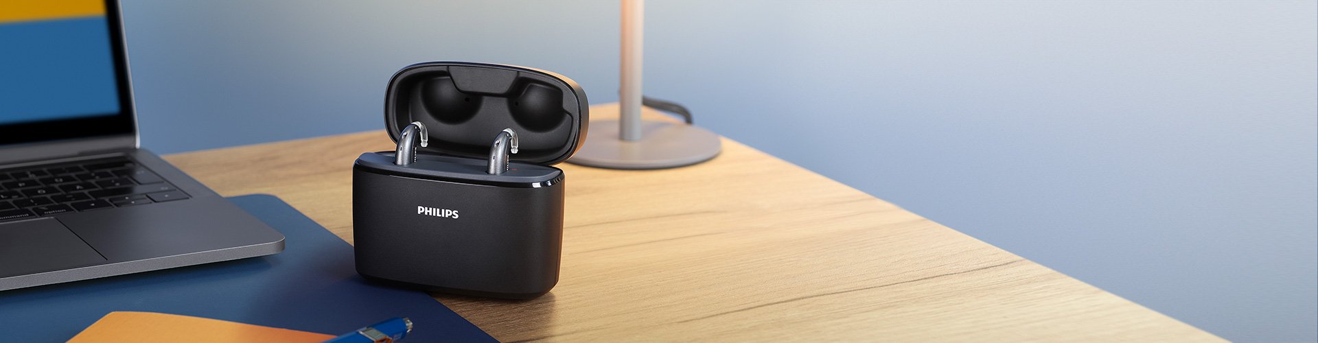 Aides auditives Philips HearLink dans le style mini contour d'oreille (miniBTE T R) sur un bureau en bois à côté d'une lampe, d'un ordinateur portable, d'un cahier et d'un stylo