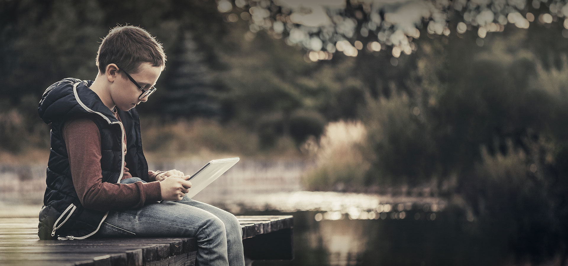 jeune garçon avec une tablette numérique assis sur un ponton de rivière