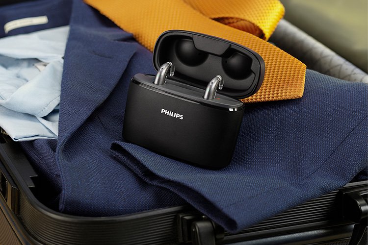 洋服が詰められたスーツケースの中で、フィリップス ヒアリンクの充電式耳かけ型補聴器 ミニBTE T Rが、 携帯用充電器チャージャープラスで充電されている