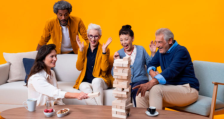 Grupa pięciu dorosłych i seniorów, w tym mężczyzna i kobieta z aparatami słuchowymi Philips HearLink, siedzą na kanapie i grają na dużym stoliku kawowym w Jengę