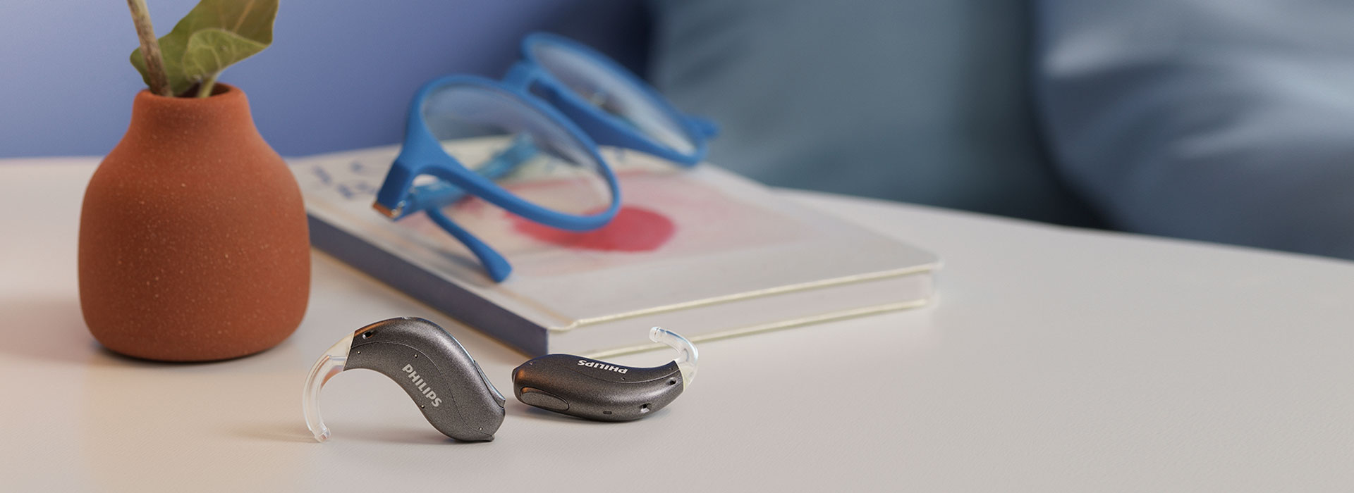 Philips HearLink hearing aids sit on a table at home next to a pair of reading glasses, a book, a lamp, and a ceramic vase with a small yellow flower